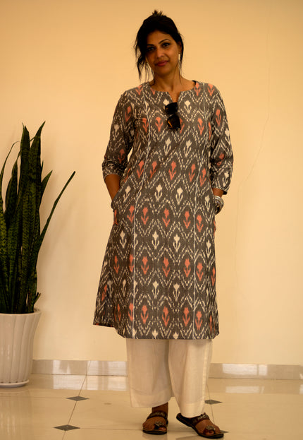 Woman wearing a patterned kurta with white pants standing indoors next to a plant.