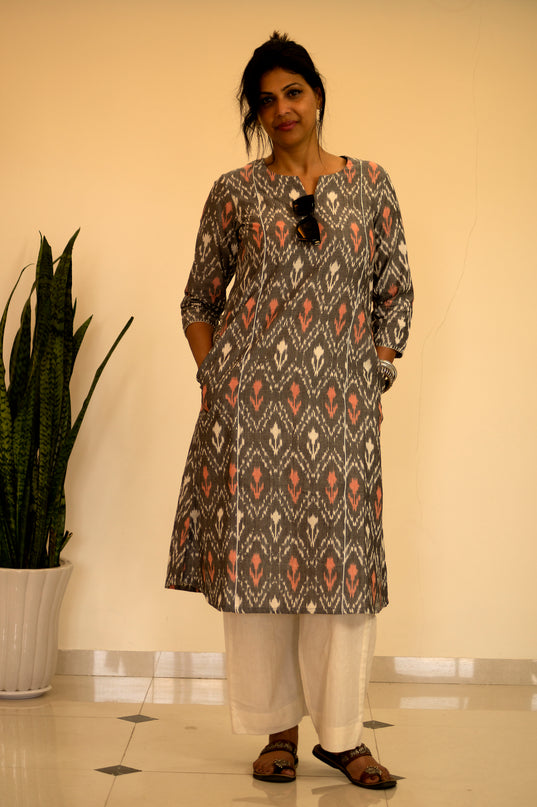 Woman wearing a patterned kurta with white pants standing indoors next to a plant.